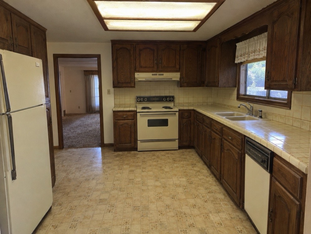 Dated 1980s kitchen with dark wood cabinets, tile counters and yellowed vinyl floor